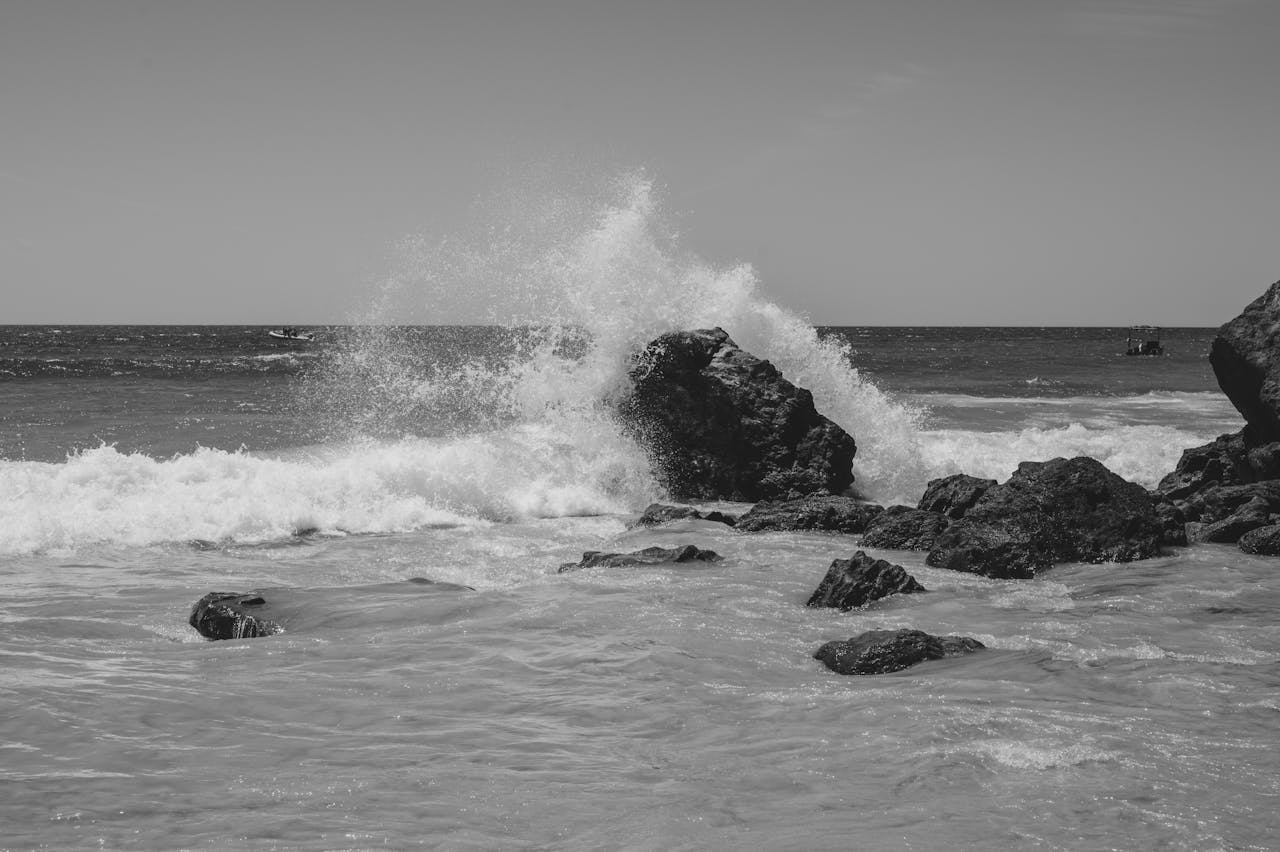 menu-01 Black and white seascape of ocean waves hitting rugged rocks under a clear sky.