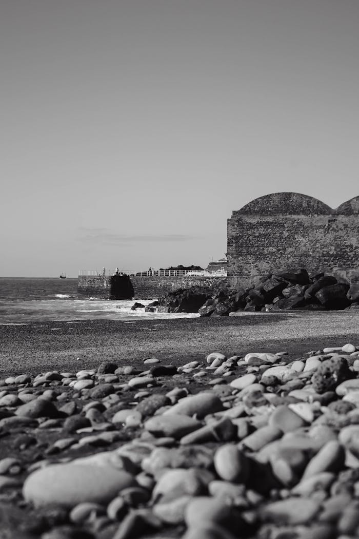about-03 Monochrome image of a rocky beach with an old stone wall by the sea under a clear sky.